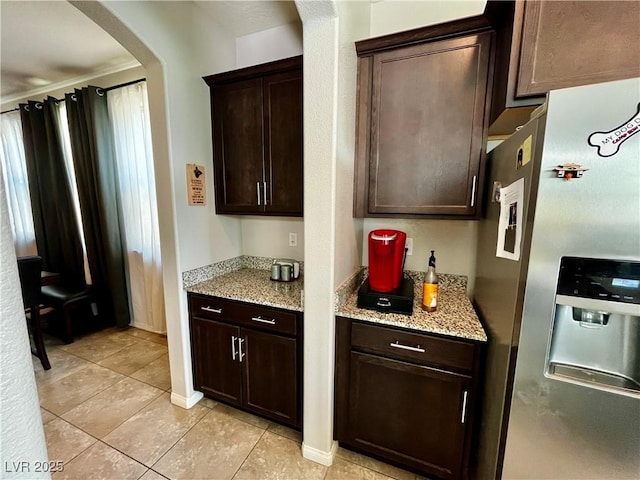 kitchen featuring stainless steel fridge with ice dispenser, dark brown cabinetry, light tile patterned floors, arched walkways, and light stone counters