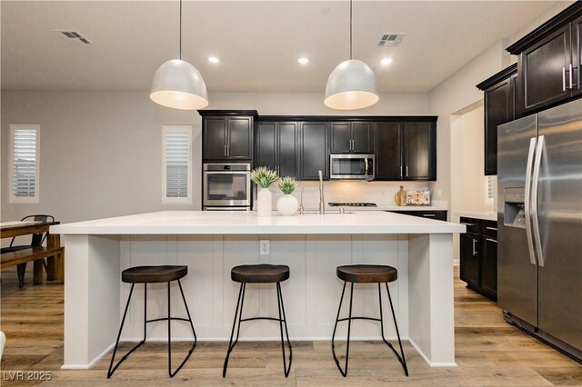 2450 Amatrice St, Henderson, NV 89044 - kitchen with stainless steel appliances, light countertops, an island with sink, decorative backsplash, and light wood-style flooring