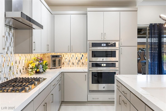 kitchen featuring appliances with stainless steel finishes, wall chimney range hood, decorative backsplash, light stone countertops, and gray cabinetry