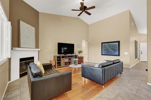 living room with a ceiling fan, high vaulted ceiling, a fireplace, and baseboards