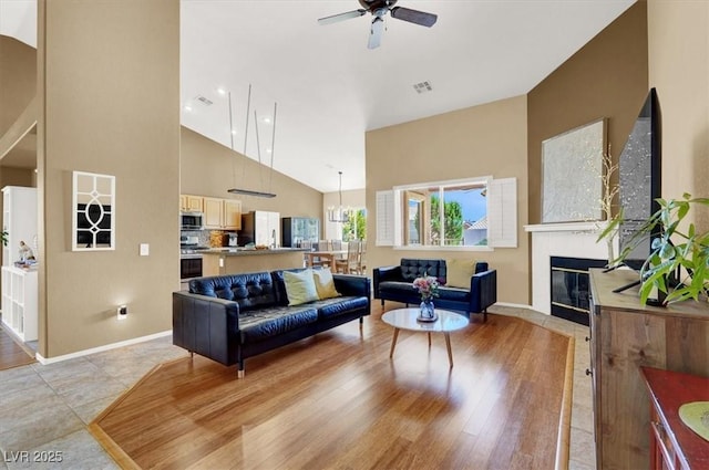 living area with a glass covered fireplace, ceiling fan, high vaulted ceiling, and baseboards
