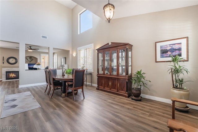 dining area featuring wood finished floors, baseboards, a ceiling fan, a glass covered fireplace, and a towering ceiling