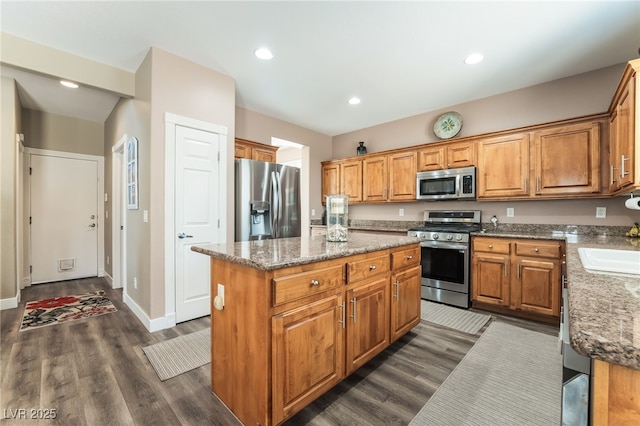 kitchen with appliances with stainless steel finishes, a kitchen island, dark wood finished floors, light stone counters, and brown cabinetry