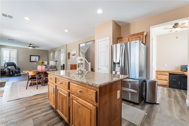 kitchen featuring stainless steel fridge with ice dispenser, a ceiling fan, a kitchen island, light stone counters, and wood finished floors