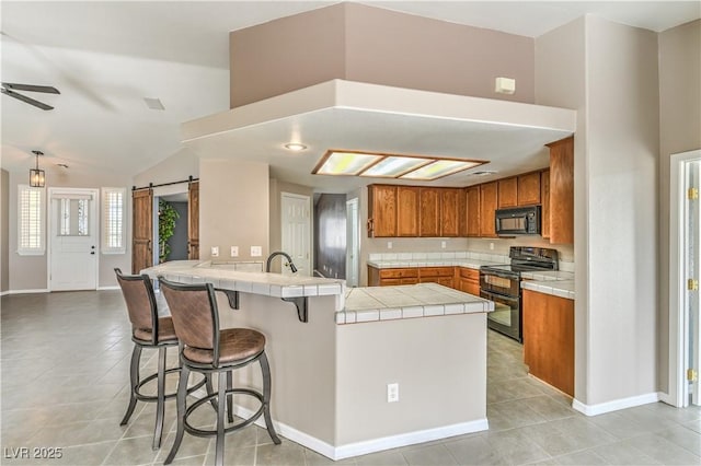 kitchen with black appliances, tile counters, brown cabinetry, a peninsula, and a barn door
