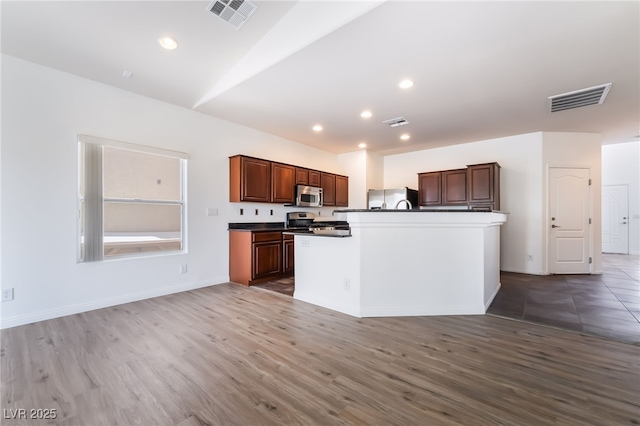 kitchen with stainless steel appliances, wood finished floors, dark countertops, recessed lighting, and a kitchen island with sink