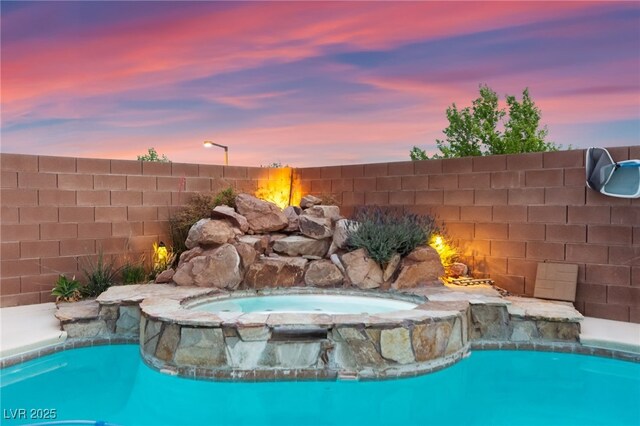 pool at dusk with fence and a pool with connected hot tub