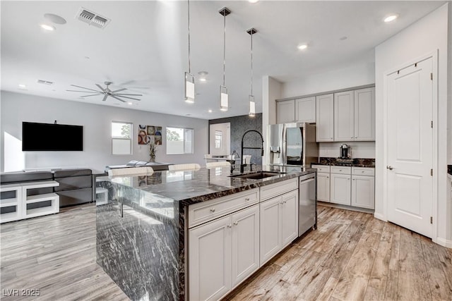 kitchen featuring a sink, stainless steel appliances, light wood-style floors, a ceiling fan, and a center island with sink
