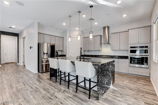 kitchen featuring stainless steel appliances, wall chimney exhaust hood, gray cabinetry, a breakfast bar area, and light wood-style flooring