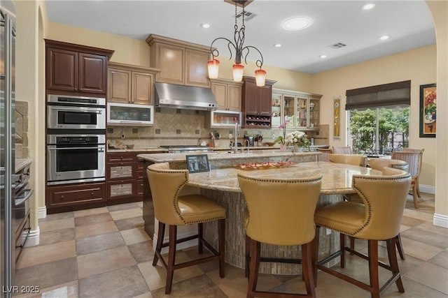 7206 Laramie Ave, Las Vegas, NV 89113 - kitchen featuring under cabinet range hood, appliances with stainless steel finishes, backsplash, light stone countertops, and an island with sink