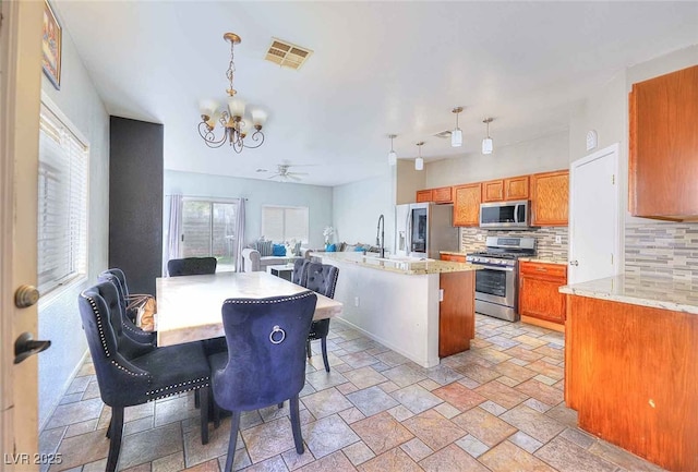 6233 Overhang Ave, Henderson, NV 89011 - kitchen with stainless steel appliances, a chandelier, decorative backsplash, brown cabinetry, and stone tile floors