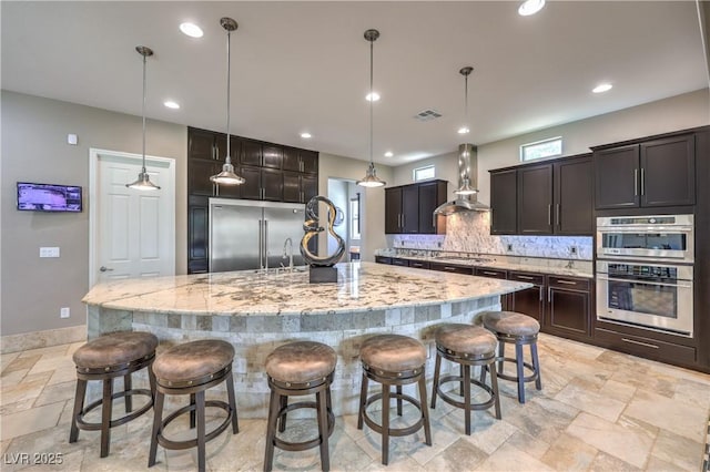 10319 Larkmead Ave, Las Vegas, NV 89149 - kitchen featuring wall chimney range hood, appliances with stainless steel finishes, tasteful backsplash, stone tile floors, and a large island