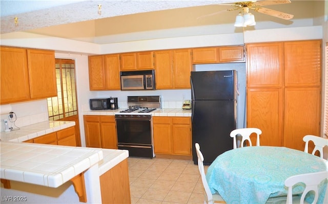 3519 W Red Coach Ave, North Las Vegas, NV 89031 - kitchen with black appliances, tile counters, a ceiling fan, light tile patterned floors, and brown cabinets