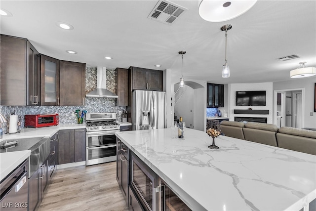 9585 Teton Diablo Ave, Las Vegas, NV 89117 - kitchen featuring wall chimney range hood, appliances with stainless steel finishes, dark brown cabinets, decorative backsplash, and hanging light fixtures
