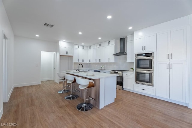 1136 Casady Hollow Ave, Henderson, NV 89012 - kitchen featuring appliances with stainless steel finishes, wall chimney exhaust hood, a breakfast bar area, light wood-style flooring, and a center island with sink