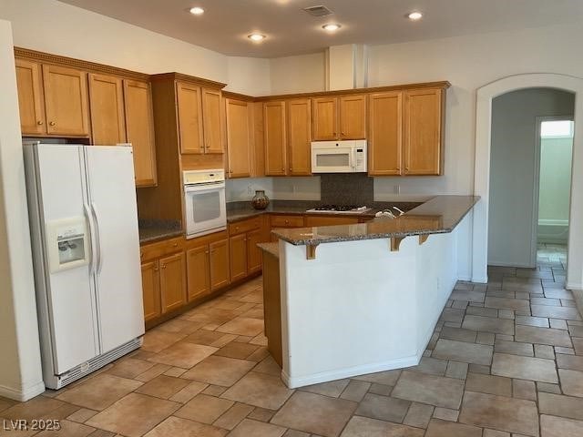 10816 Elm Ridge Ave, Las Vegas, NV 89144 - kitchen featuring white appliances, arched walkways, a peninsula, dark stone countertops, and a kitchen bar