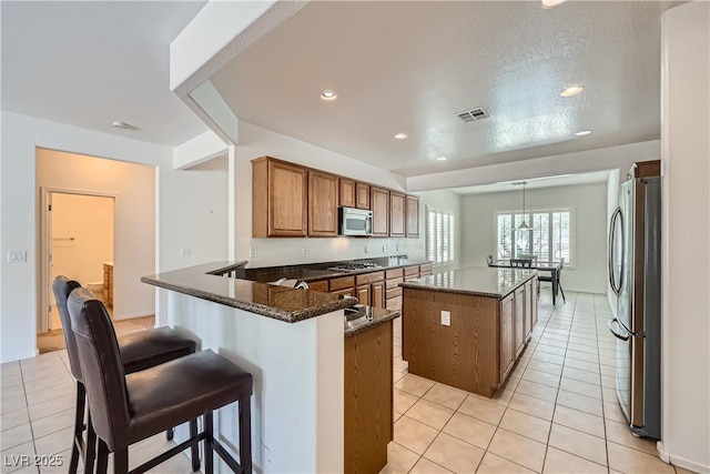 7465 Widewing Dr, North Las Vegas, NV 89084 - kitchen featuring appliances with stainless steel finishes, brown cabinets, a center island, light tile patterned flooring, and dark stone counters