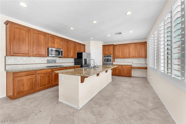1155 Phillip Island St, Henderson, NV 89052 - kitchen with stainless steel appliances, decorative backsplash, brown cabinetry, light stone counters, and a center island with sink