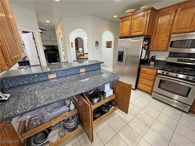 3451 Sagebrush Ave, Pahrump, NV 89048 - kitchen with appliances with stainless steel finishes, arched walkways, light tile patterned floors, brown cabinetry, and dark stone countertops