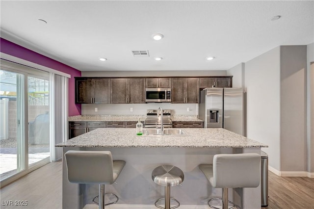 6940 Crescent Gold St, North Las Vegas, NV 89086 - kitchen with appliances with stainless steel finishes, dark brown cabinets, light wood-style flooring, a breakfast bar, and a kitchen island with sink