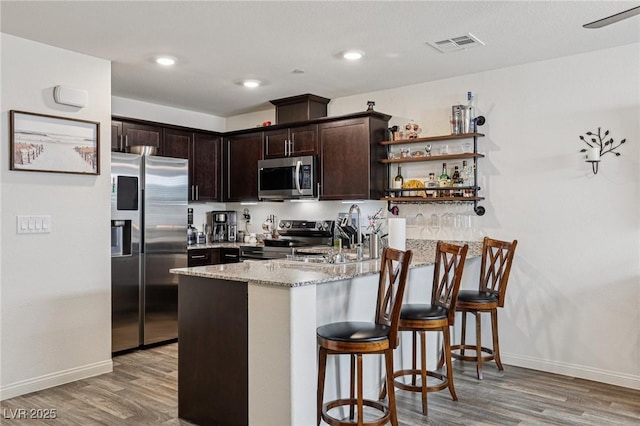 6807 Satin Silver Ct, North Las Vegas, NV 89086 - kitchen with stainless steel appliances, a peninsula, dark brown cabinetry, light stone counters, and light wood-style flooring