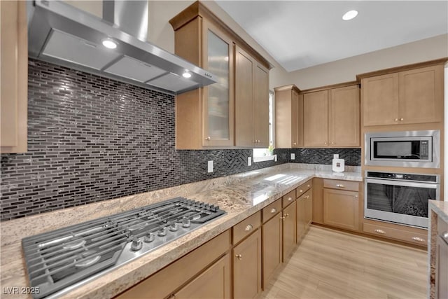 7258 W Le Baron Ave, Las Vegas, NV 89178 - kitchen with appliances with stainless steel finishes, wall chimney range hood, decorative backsplash, glass insert cabinets, and light wood-style flooring