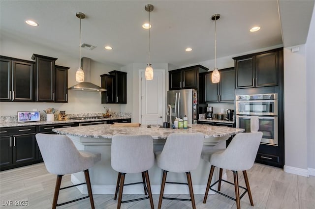 7194 Sage Wren Ct, Las Vegas, NV 89148 - kitchen featuring wall chimney exhaust hood, stainless steel appliances, a breakfast bar, a center island with sink, and light stone countertops