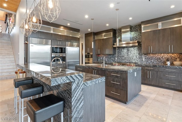 8 Meadowhawk Ln, Las Vegas, NV 89135 - kitchen with a kitchen island with sink, wall chimney exhaust hood, built in appliances, decorative backsplash, and dark brown cabinets