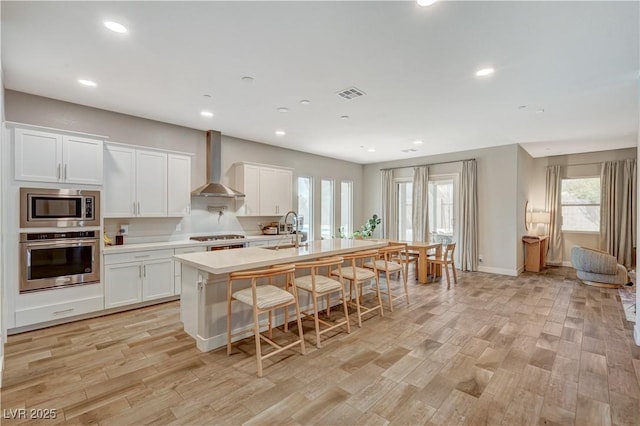 7736 Tioga Ridge Ct, Las Vegas, NV 89117 - kitchen with stainless steel appliances, wall chimney range hood, an island with sink, light countertops, and light wood-type flooring