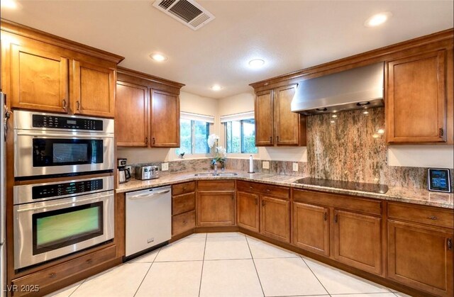 3597 Cochise Ln, Las Vegas, NV 89169 - kitchen with appliances with stainless steel finishes, wall chimney exhaust hood, brown cabinetry, light tile patterned flooring, and tasteful backsplash