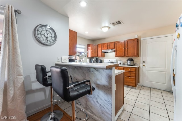 4656 E Desert Inn Rd, Las Vegas, NV 89121 - kitchen with under cabinet range hood, brown cabinetry, light countertops, light tile patterned floors, and a kitchen breakfast bar