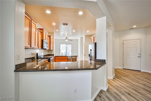 5967 Sun Appello Ave, Las Vegas, NV 89122 - kitchen featuring stainless steel appliances, a peninsula, light wood-style flooring, brown cabinetry, and dark stone countertops
