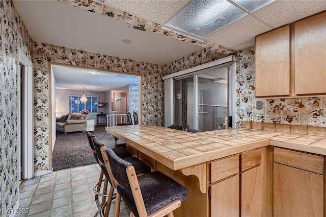 6213 Fairwood Ave, Las Vegas, NV 89107 - kitchen featuring wallpapered walls, tile countertops, a peninsula, light brown cabinets, and open floor plan
