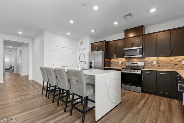 5936 Bruselas St, Las Vegas, NV 89113 - kitchen featuring appliances with stainless steel finishes, an island with sink, backsplash, light stone countertops, and light wood-style flooring