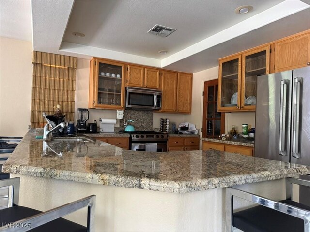 8225 Bermuda Beach Dr, Las Vegas, NV 89128 - kitchen featuring stainless steel appliances, a peninsula, brown cabinets, a kitchen bar, and glass insert cabinets
