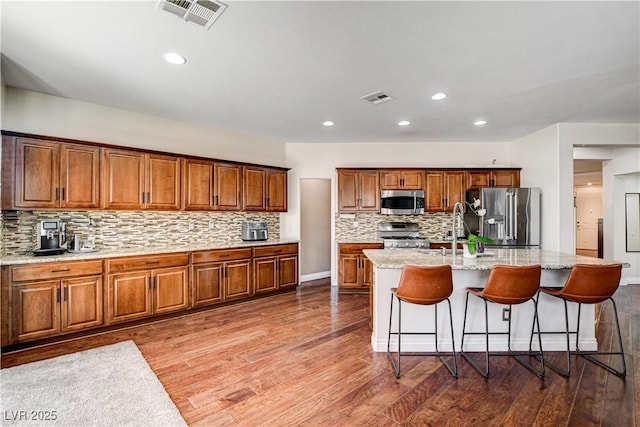 1717 Jake Andrew Ave, North Las Vegas, NV 89086 - kitchen featuring stainless steel appliances, brown cabinetry, backsplash, a center island with sink, and wood finished floors