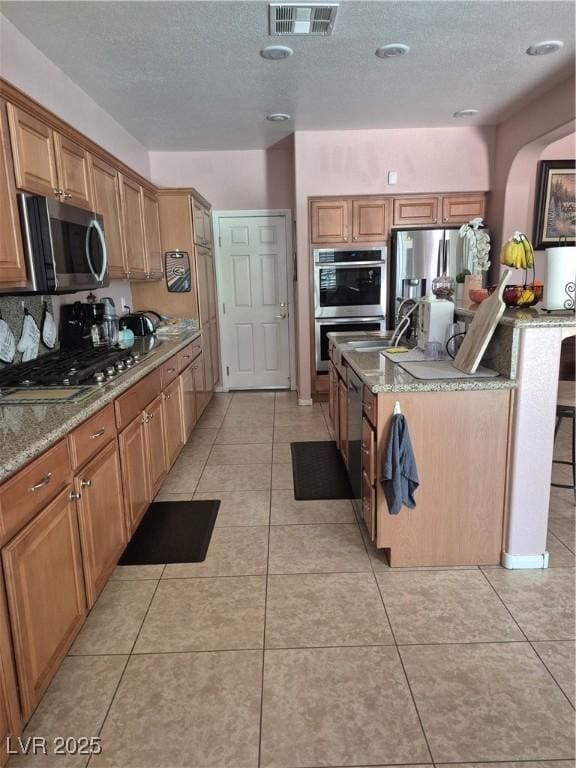 6009 Arbor Crest St, North Las Vegas, NV 89081 - kitchen featuring stainless steel appliances, light tile patterned floors, a textured ceiling, light stone counters, and brown cabinetry