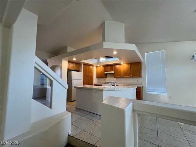8704 Autumn Wreath Ave, Las Vegas, NV 89129 - kitchen with freestanding refrigerator, under cabinet range hood, brown cabinetry, tile countertops, and a center island