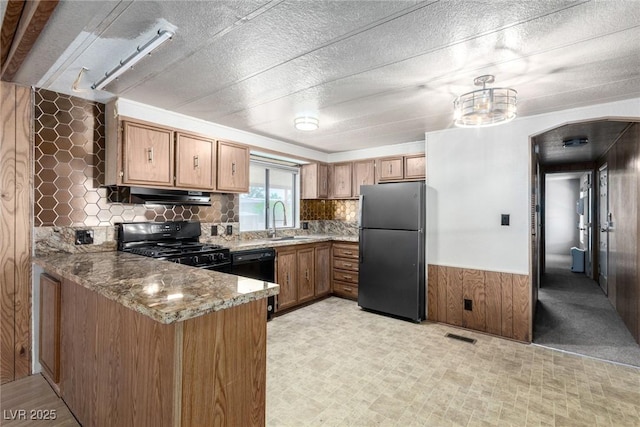 3471 Cape Cod Dr, Las Vegas, NV 89122 - kitchen with black appliances, a peninsula, brown cabinets, under cabinet range hood, and light stone counters