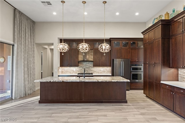 10179 Emerald Sunset Ct, Las Vegas, NV 89148 - kitchen with appliances with stainless steel finishes, wall chimney range hood, decorative backsplash, an island with sink, and dark brown cabinets