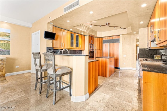 2877 Paradise Rd Unit 702, Las Vegas, NV 89109 - kitchen with brown cabinetry, built in appliances, decorative backsplash, a peninsula, and dark stone counters