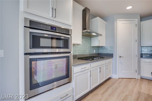 10342 Kesington Dr, Las Vegas, NV 89135 - kitchen featuring appliances with stainless steel finishes, wall chimney exhaust hood, white cabinets, light wood-style flooring, and tasteful backsplash