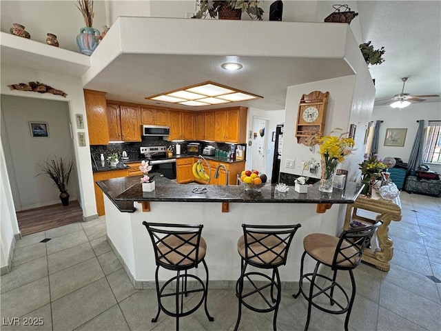 4910 Marywood Ct, Pahrump, NV 89061 - kitchen with stainless steel appliances, a peninsula, brown cabinetry, light tile patterned flooring, and tasteful backsplash