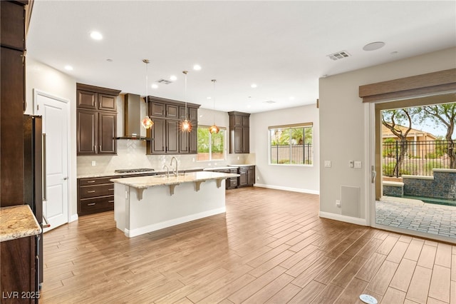 2116 Canvas Edge Dr, Henderson, NV 89044 - kitchen with wall chimney exhaust hood, a kitchen island with sink, dark brown cabinetry, decorative backsplash, and light wood finished floors