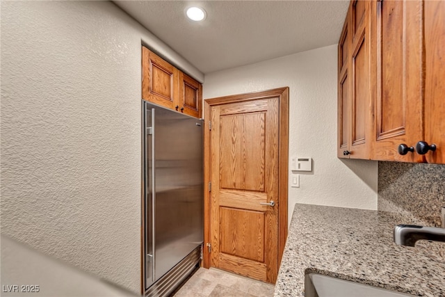 7045 Edna Ave, Las Vegas, NV 89117 - kitchen featuring built in fridge, a textured wall, brown cabinetry, light stone counters, and recessed lighting
