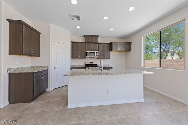 3912 Makushin Bay Ave, North Las Vegas, NV 89081 - kitchen featuring appliances with stainless steel finishes, dark brown cabinets, light stone countertops, a center island with sink, and recessed lighting
