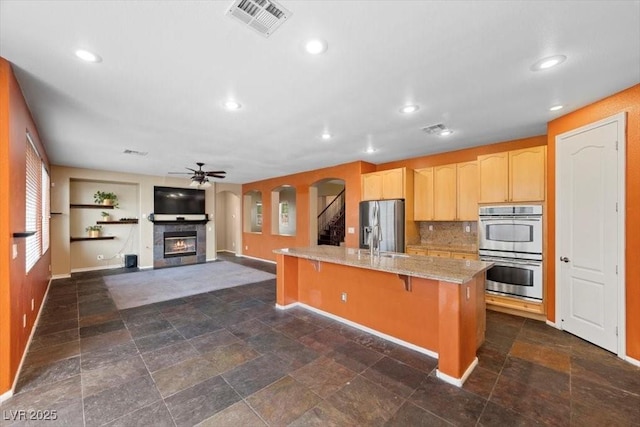 9413 Arcadia Woods Ct, Las Vegas, NV 89149 - kitchen with stainless steel appliances, a glass covered fireplace, light brown cabinets, a ceiling fan, and a breakfast bar area