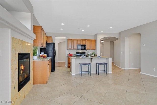 7558 Schuders Ave, Las Vegas, NV 89178 - kitchen featuring black appliances, arched walkways, light tile patterned floors, a center island with sink, and a premium fireplace
