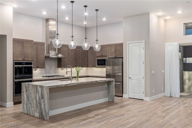 4152 Yucca Bloom St Unit 1, Las Vegas, NV 89135 - kitchen featuring stainless steel appliances, wall chimney range hood, light wood-style flooring, backsplash, and a center island with sink