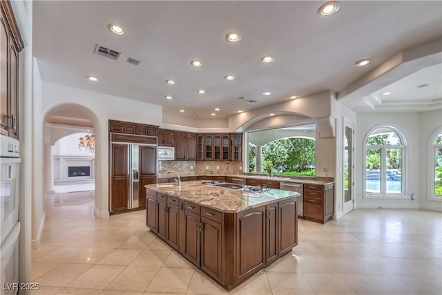 8021 Obannon Dr, Las Vegas, NV 89117 - kitchen featuring built in appliances, arched walkways, a center island with sink, tasteful backsplash, and light tile patterned floors